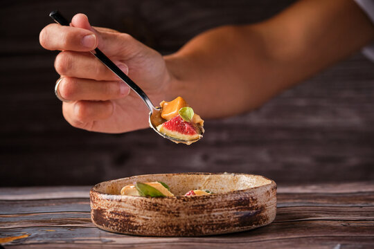 Cropped unrecognizable person with spoon of tasty caramel cheesecake with shipped cream and piece of fig at wooden table in cafe