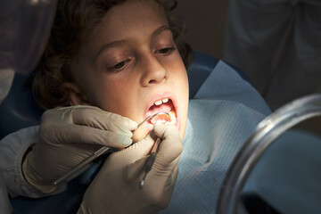 From above of unrecognizable dentist in protective costume and gloves examining teeth of child lying in dental chair during coronavirus pandemic