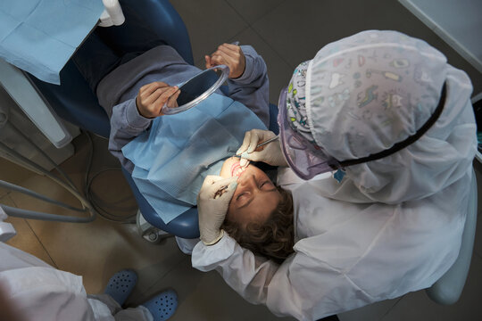 From above of unrecognizable dentist in protective costume and gloves examining teeth of child lying in dental chair during coronavirus pandemic