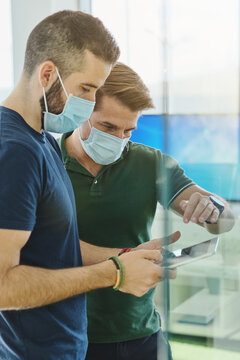 Through glass busy male programmers in medical masks standing with tablet in office and discussing software development at work during coronavirus outbreak