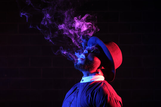 Side view of stylish African American male in hat and sunglasses standing on black background in studio with neon illumination and smoking vape while exhaling steam