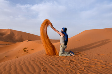Kneeling occidental man throwing silky orange sand from the top of a sand dune in the desert, creating a sandy orange flame shape in the air