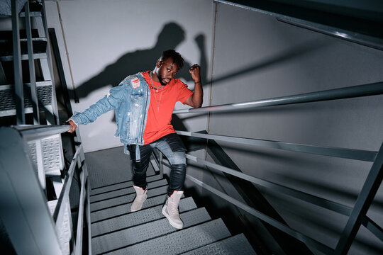 High angle of thoughtful African American male in trendy apparel and with denim jacket standing on metal staircase and looking at camera