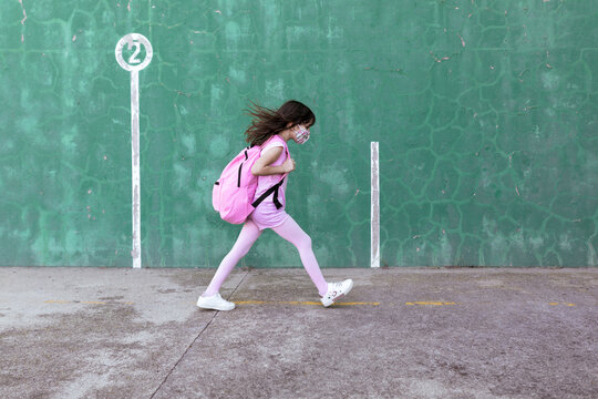 Side view of schoolgirl with rucksack and in protective mask walking along street during coronavirus epidemic