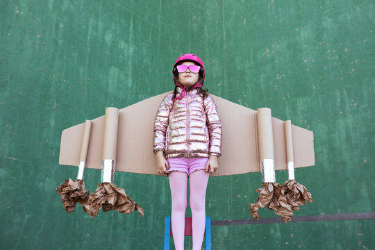 Cute girl in pilot helmet and handmade creative carton wings standing on street on green background looking up