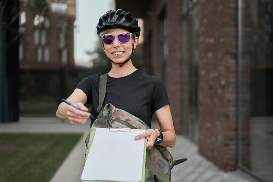 Happy woman cyclist courier with pen in hands on street on summer day