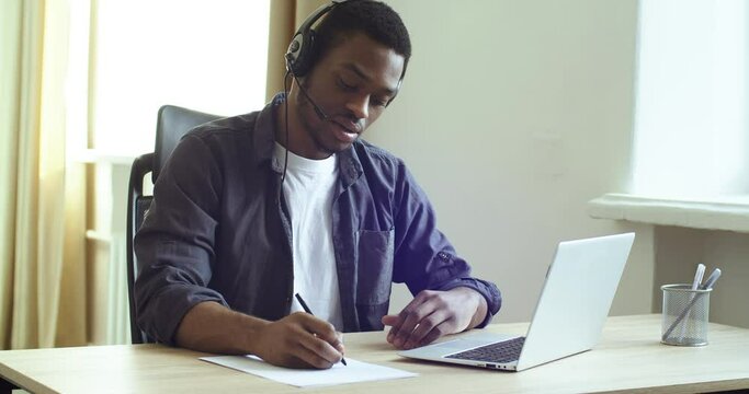 Handsome afro american man black guy in headphones student teacher speaks online remotely via video call chat conference studying remotely writes notes with pen on paper, work study during quarantine