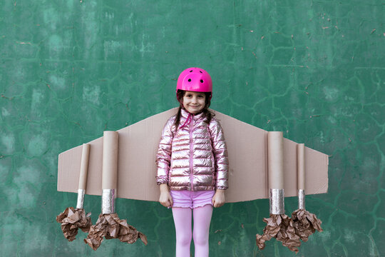 Cute girl in pilot helmet and handmade creative carton wings standing on street on green background looking at camera