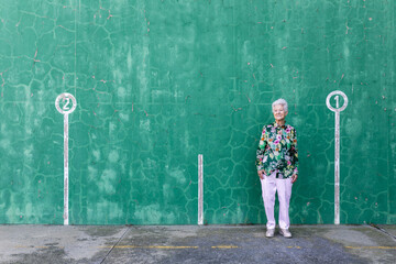 Full body of elegant elderly female in stylish outfit standing against green wall with numbers on parking lot