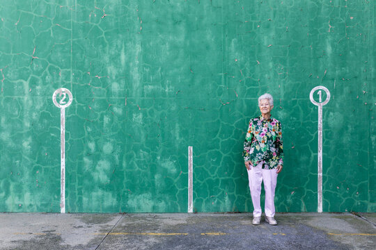Full body of elegant elderly female in stylish outfit standing against green wall with numbers on parking lot