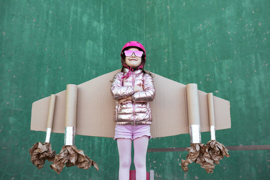 Cute girl in pilot helmet and handmade creative carton wings standing on street on green background looking up