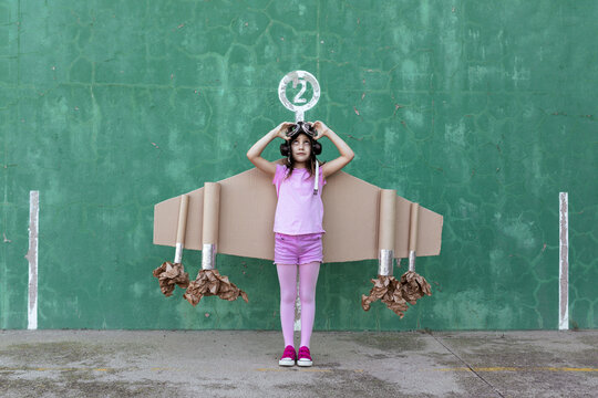 Cute girl in pilot helmet and handmade creative carton wings standing on street with raised arms looking up