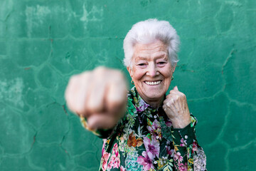 Cheerful elderly gray haired lady in stylish colorful blouse having fun and boxing at camera while standing against green wall