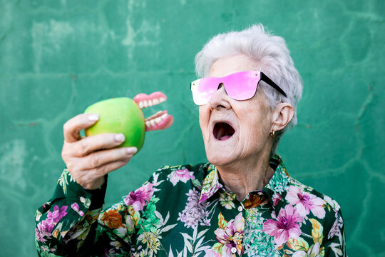Stylish elderly female in trendy outfit and sunglasses holding green apple with denture while standing against green background