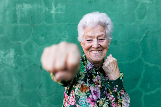 Cheerful Elderly Gray Haired Lady In Stylish Colorful Blouse Having Fun And Boxing At Camera While Standing Against Green Wall