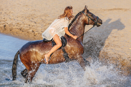 Beautiful woman in a white dress riding a horse crossing a river at sunset