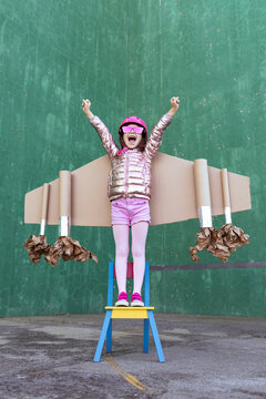 Positive girl in cardboard plane wings and helmet standing on chair with fists up while celebrating achievement and screaming on green background