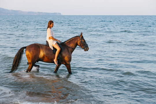 Beautiful woman in white dress riding a horse along the seashore at sunset