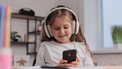 Portrait of happy little cute girl wearing headphones,choosing favorite music tracks in mobile application,texting messages,browsing internet,watching video, playing online game on smartphone at home.