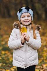 A beautiful cute girl, a child in a white jacket of preschool age, stands and poses with a yellow leaf in nature. Autumn portrait, photography.