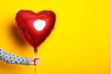 Female hand holds an inflatable heart-shaped balloon on a yellow background.
