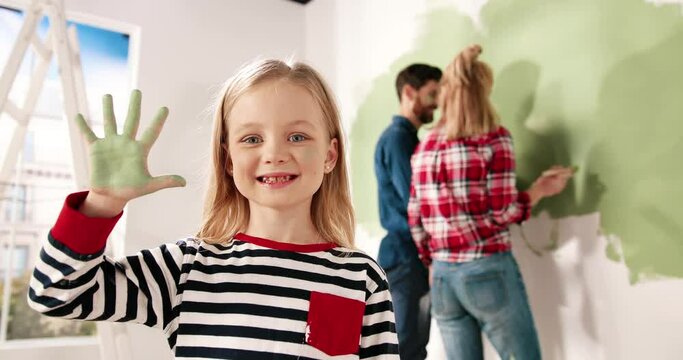 Close up portrait of joyful little cute girl smiling to camera showing hand palm in green paint. Mom and dad painting wall on background. Home repair and remodeling. Renewing house. Interior design