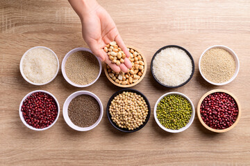 Various cereal grain in a bowl with hand on wooden background (quinoa seeds, peanut, perilla seeds, soybean, azuki beans, rice grain and mung beans)
