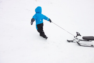 a boy in a blue winter suit pulls a snow scooter up the mountain. concept of winter recreation on the slides.