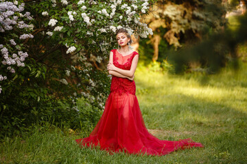 Obraz na płótnie Canvas Beautiful gorgeous woman in long red dress in blooming lilac garden. Growth Portrait in sunset light. Pretty young caucasian girl walks in garden and plays with her dress. Spring time. Young queen