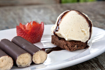 Chocolate covered walnut ice cream rolls, ice cream scoop and cake with a strawberry served on a white plate on wooden table