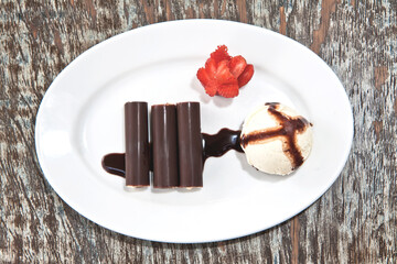 Chocolate covered walnut ice cream rolls, ice cream scoop and cake with a strawberry served on a white plate on wooden table