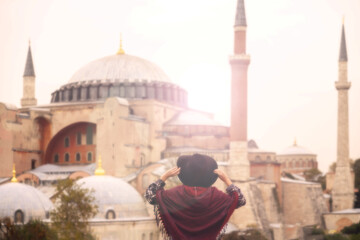 Fototapeta premium A young girl in a red poncho holds a hat and looks at the Hagia Sophia mosque. Istanbul, Turkey.