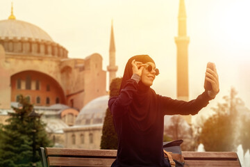 A young smiling Muslim girl in a hijab and stylish black glasses makes a selfie against the background of the Hagia Sophia mosque. Tourism in Istanbul, Turkey