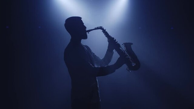 Backlit silhouette of saxophonist man with saxophone standing in dark nightclub disco studio and start playing sax. Light appears and illuminates the man. Isolated on black background. Slow motion