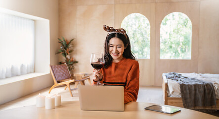 Portrait of beautiful business asian woman online working in bedroom desk use computer. Freelance online marketing, girl video call on laptop toasting with wine celebrating Christmas alone at home.