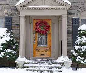 Front door of house with colorful Christmas wreath and snow covered evergreen bushes