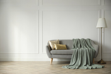 Stylish room interior with comfortable sofa, knitted blanket and lamp near white wall. Space for text