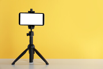 Smartphone with blank screen fixed to tripod on white table against yellow background. Space for text