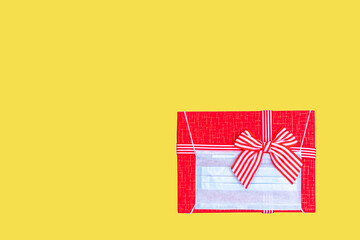 Gift concept during the coronavirus pandemic. Red gift box with wearing a protective medical mask on trendy Illuminating yellow 2021 background, copy space