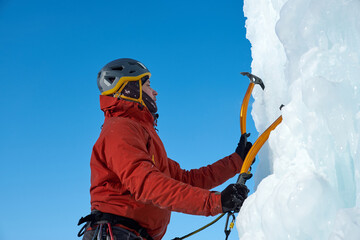 Alpinist man with ice tools axe climbing a large wall of ice. Outdoor Sports Portrait.