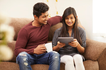 Cheerful young beautiful couple chatting with tablet and coffee on sofa at home.