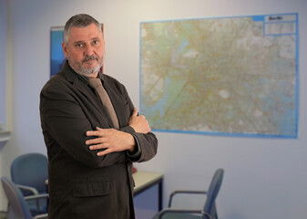 An elderly man with a beard stands in an office with his arms crossed. He looks skeptically into the camera. In the background there is a city map on the wall and there are office chairs.
