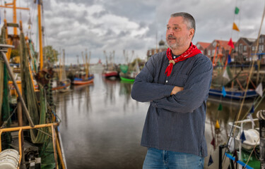 In front of the small port of Neuharlingersiel stands a man with a beard in a typical fisherman's shirt with a red scarf. In the background are the boats of the shrimp fishermen.