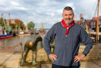 A man with a beard in a typical fisherman's shirt with a red scarf stands in front of the small port of Neuharlingersiel in northern Germany with fishing boats.