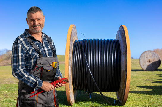 A bearded worker in work trousers stands in front of a large cable reel with pliers in hand. In the background is another cable reel on a meadow in the sunshine.