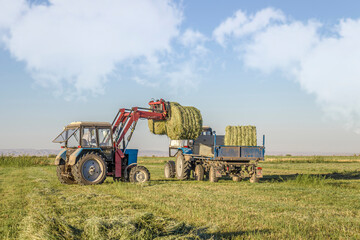 Obraz premium Bales of hay are loaded onto a trailer by a tractor in the countryside during the summer season