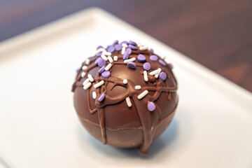 Close up food photograph of a hot cocoa or chocolate bomb covered in drizzled brown chocolate and purple and white sprinkles set on a rectangular plate making a great gift during the winter holidays.