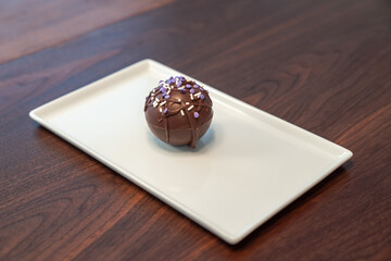 Food photograph of a single hot cocoa or chocolate bomb covered in drizzled brown chocolate and purple and white sprinkles set on a white serving plate making a great gift during the winter holidays.
