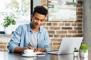 happy african american man writing in notebook near laptop and cup on desk.