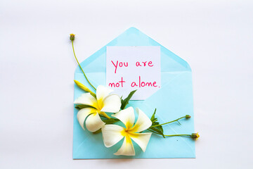 you are not alone message card handwriting in blue envelope with white flowers fragnipani arrangement flat lay postcard style on background white